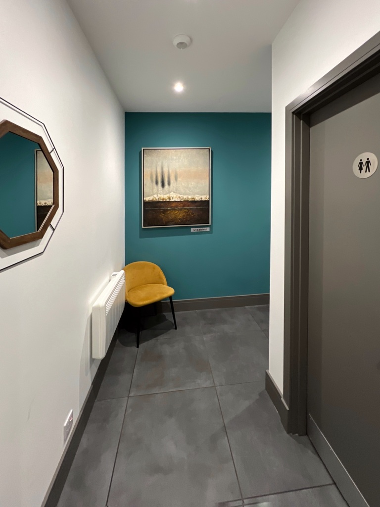 The hallway to the cludgies, with a white wall to the left (with a mirror on it), and a turquoise wall at the back with a large painting hanging on it. To the right is a door to the unisex cludgie.